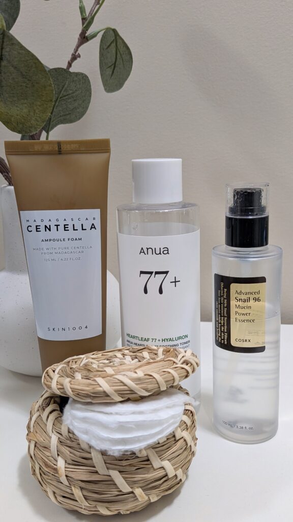 A lineup of the SKIN1004 Centella foam cleanser, Anua Heartleaf 77 toner, and COSRX snail mucin essence on a vanity.
